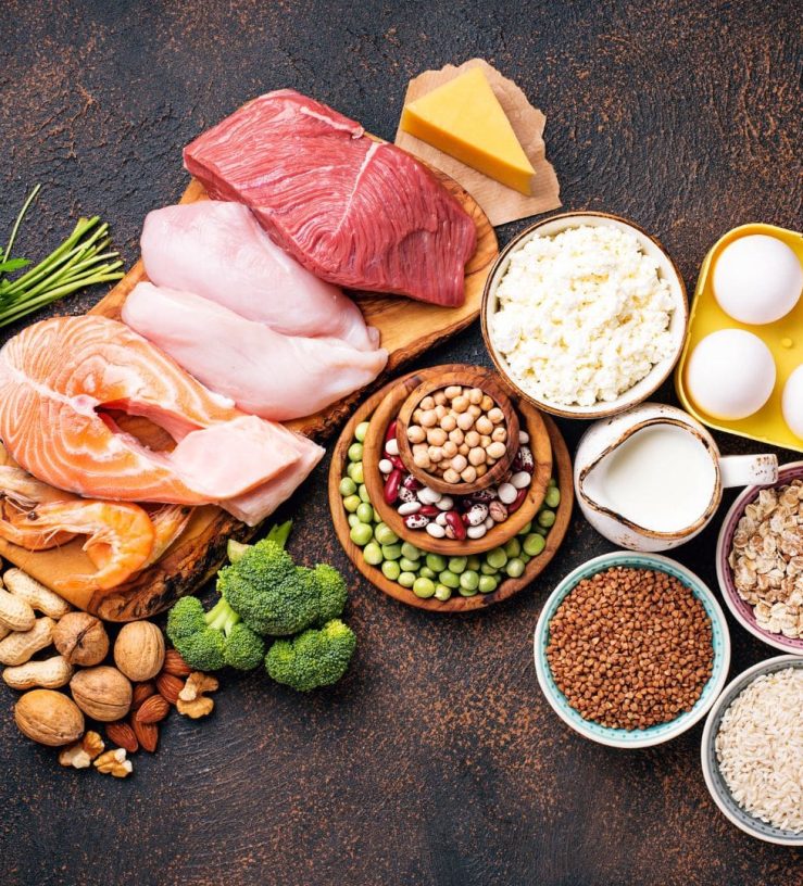 A photo of several foods on a table which are all high in protein such as meat, salmon, eggs and nuts.