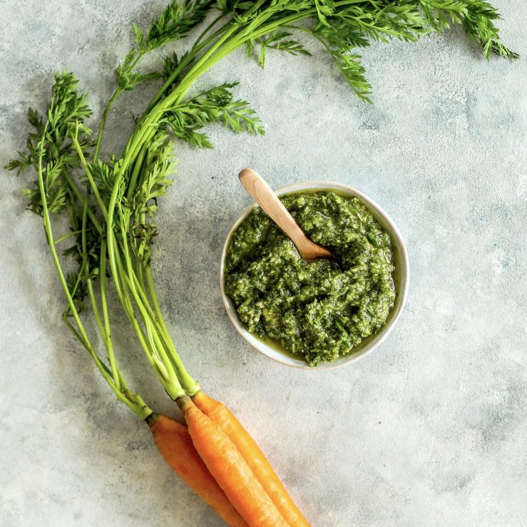 Pot of green carrot top pesto, shown on bench top with small bunch of fresh carrots and leafy green carrot tops