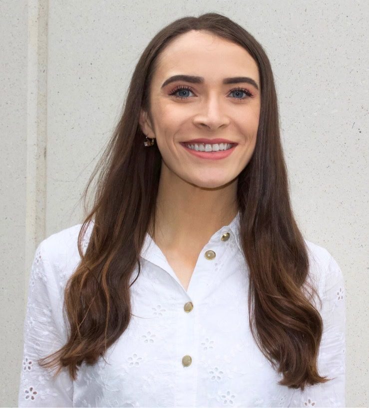 A photo of Estelle smiling, she has long, brown hair and blue eyes and is wearing a white shirt