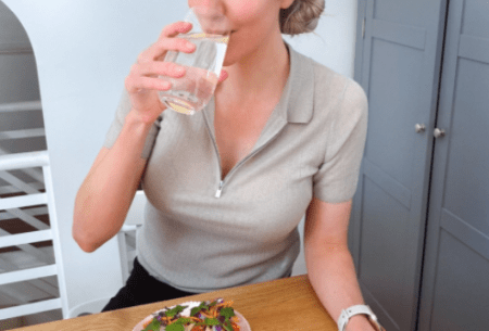 Dr Megan Rossi sitting at a table drinking a glass of water with a small plate of snacks in front of her